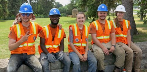 Northern Bedrock team Andrew, Gana, Sunflower, Michael, and Naomi, sittin on a wall at Graeser Park with the beehive in the background.